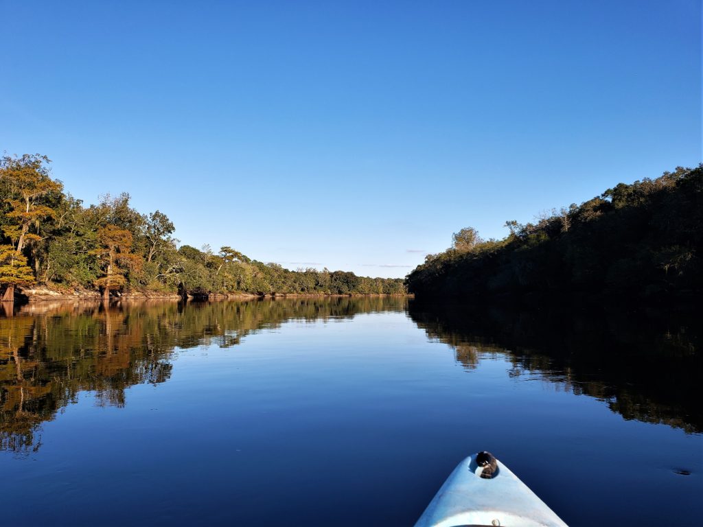 The front edge of a kayak with the blue sky and water of the river and the trees reflecting in the glassy water