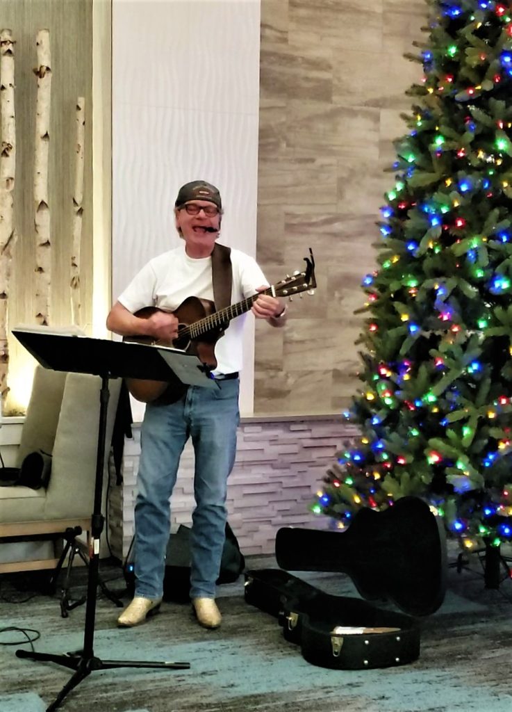 Man playing guitar and singing, next to a christmas tree