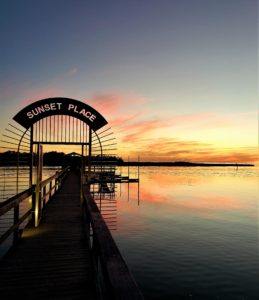 A long dock on the Steinhatchee River with sign over it called Sunset Place, The sky is pink, orange, brown and blue and reflecting in the water.