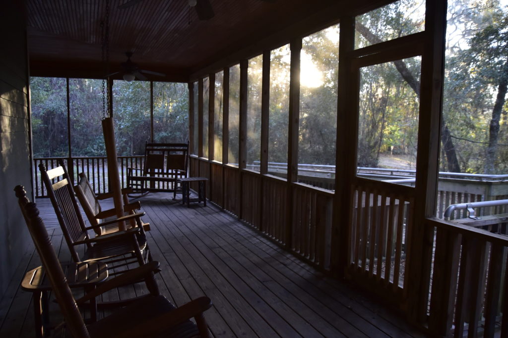 Screened back porch of cabin with rockers and swing, surrounded by trees and sun setting